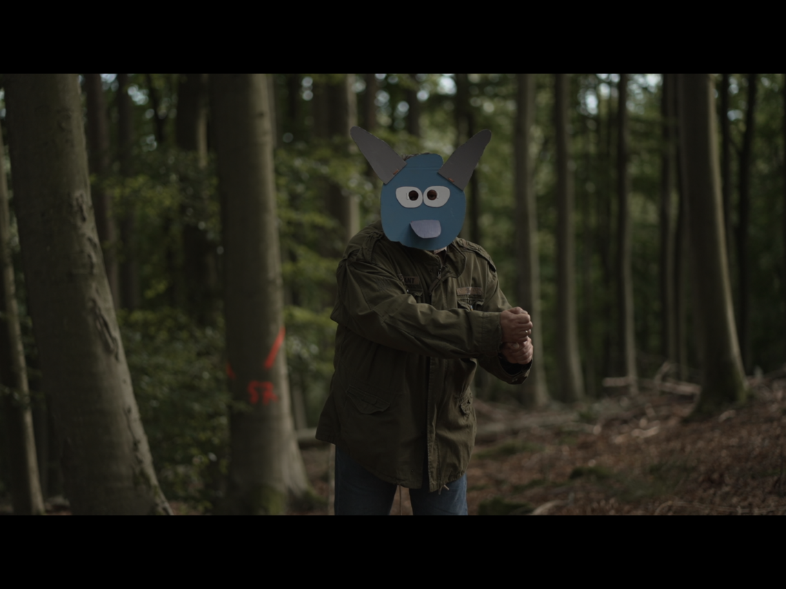 Eine Person steht im Wald und gestikuliert, sie trägt eine bunte Maske aus Pappe, die ein Fabelwesen darstellt.  | © Mia & Eric