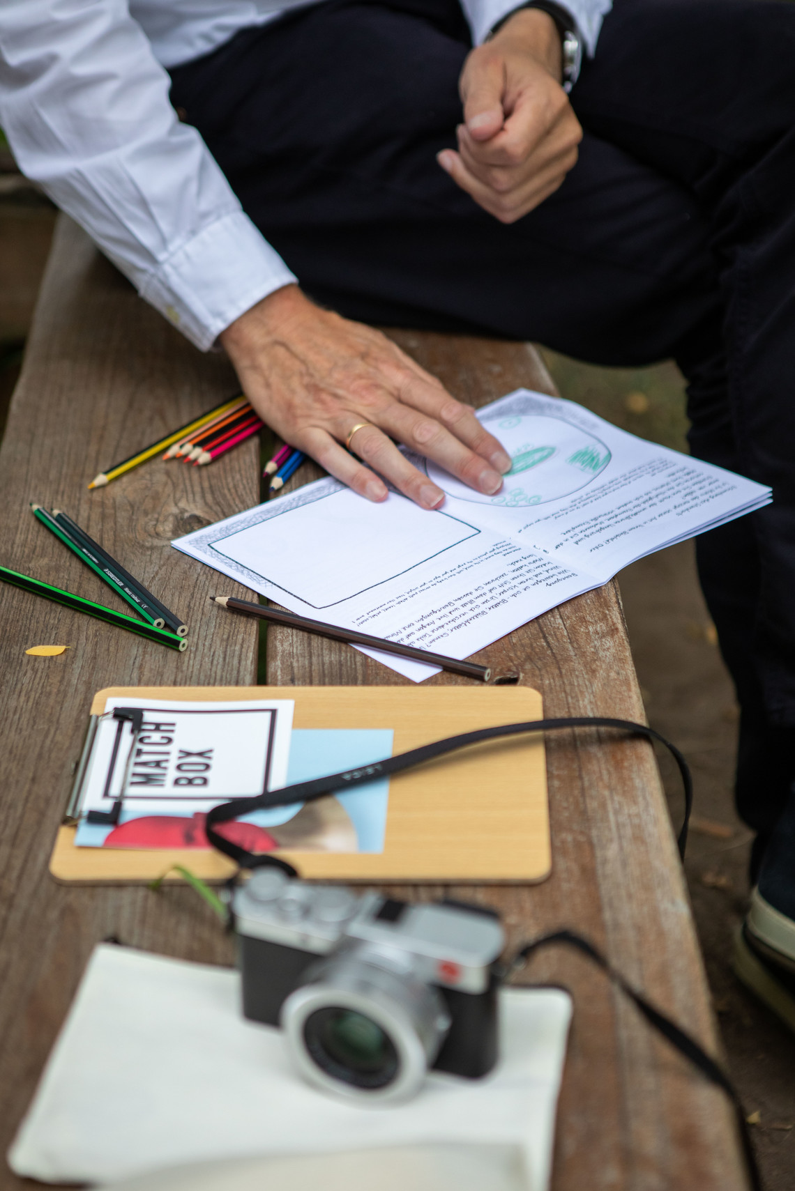 Ein Mann sitzt auf einer Bank mit Materialien: Sticker, Kamera, Stifte | © Arthur Bauer