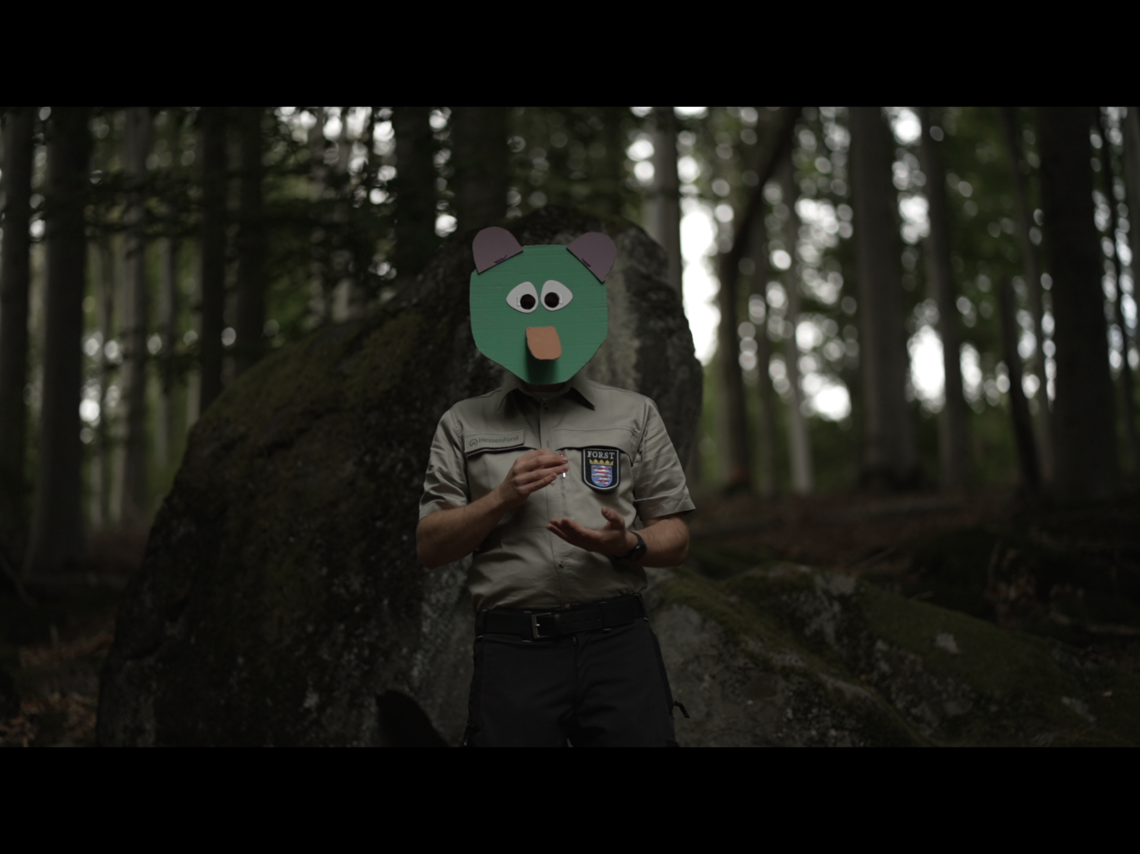 Eine Person steht im Wald und gestikuliert, sie trägt eine bunte Maske aus Pappe, die ein Fabelwesen darstellt.  | © Mia & Eric