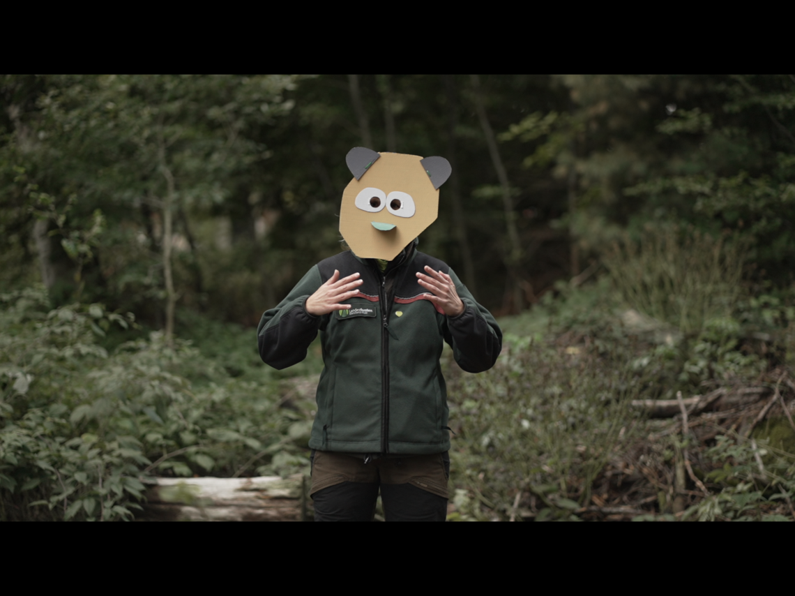 Eine Person steht im Wald und gestikuliert, sie trägt eine bunte Maske aus Pappe, die ein Fabelwesen darstellt.  | © Mia & Eric