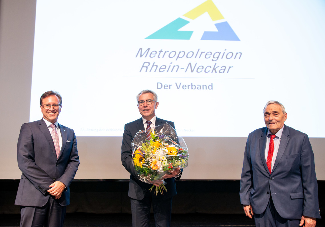 drei Personen stehen auf der Bühne, eine davon mit Blumen in der Hand  | © VRRN e.v.