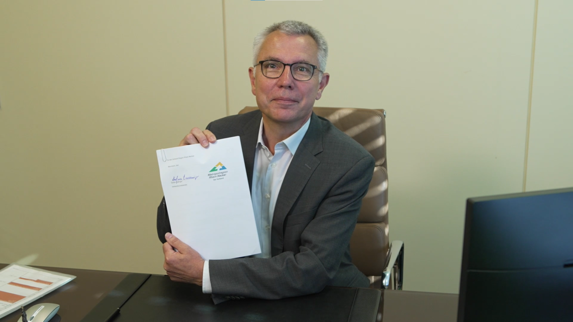 Stefan Dallinger sitzt an seinem Schreibtisch und hält den unterschriebenen Mobilitätspakt Rhein-Neckar in die Kamera | © RNK