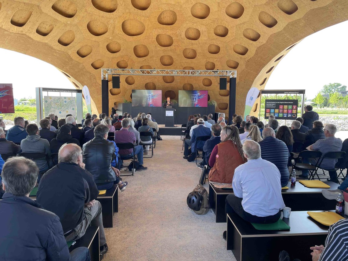 Menschen sitzen vor einer Bühne am Holzpavillon der Region auf der BUGA in Mannheim | © VRRN/Lersch