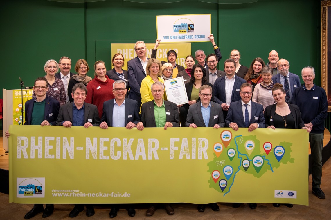 Menschen stehen hinter einem Banner von Rhein-Neckar Fair und lächeln. Hinten halten zwei Männer ein Schild hoch mit der Aufschrift: "Wir sind Faire Region" | © VRRN / Schwerdt