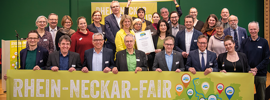Menschen stehen hinter einem Banner von Rhein-Neckar Fair und lächeln. Hinten halten zwei Männer ein Schild hoch mit der Aufschrift: "Wir sind Faire Region" | © VRRN / Schwerdt