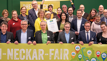 Menschen stehen hinter einem Banner von Rhein-Neckar Fair und lächeln. Hinten halten zwei Männer ein Schild hoch mit der Aufschrift: "Wir sind Faire Region" | © VRRN / Schwerdt