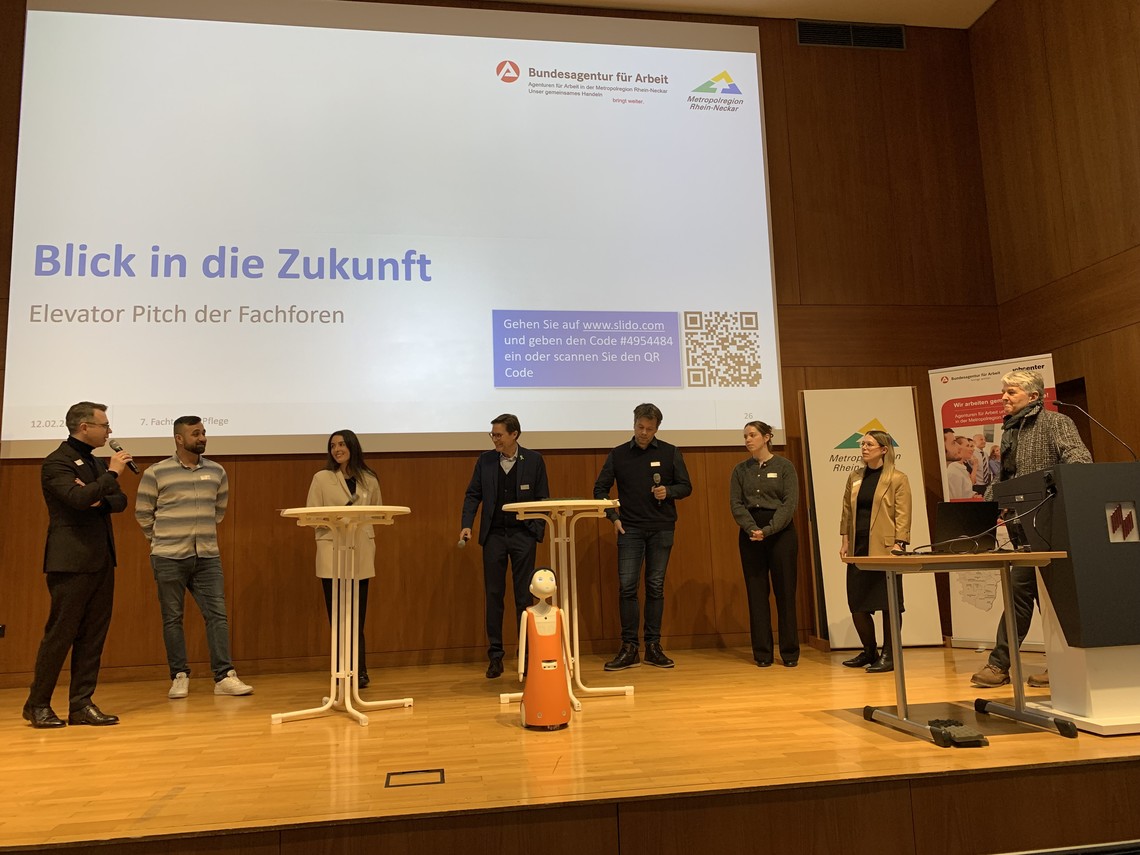 Menschen auf einer Bühne an Stehtischen und ein kleiner Roboter davor. | © VRRN / LH