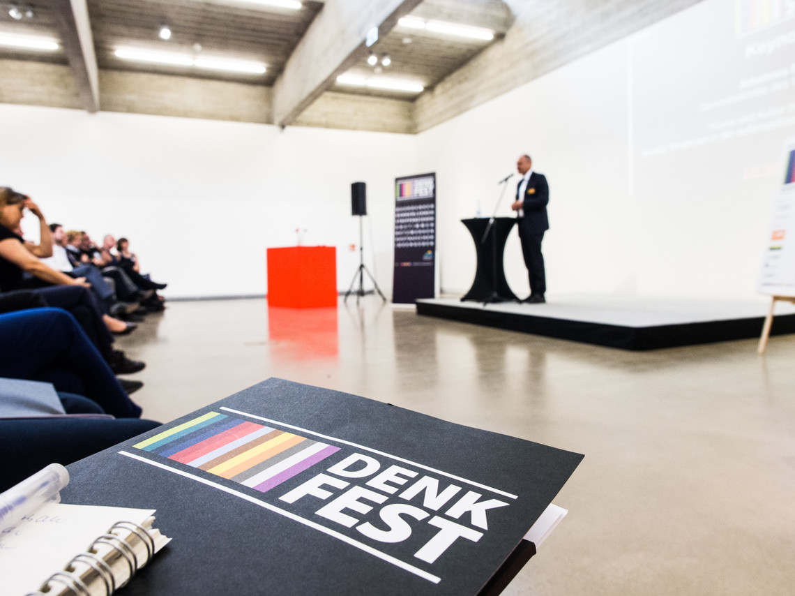 Eine Mappe mit Denkfest-Logo im Fokus, im Hintergrund steht ein Mensch auf einer Bühne | © Gunnar Fuchs