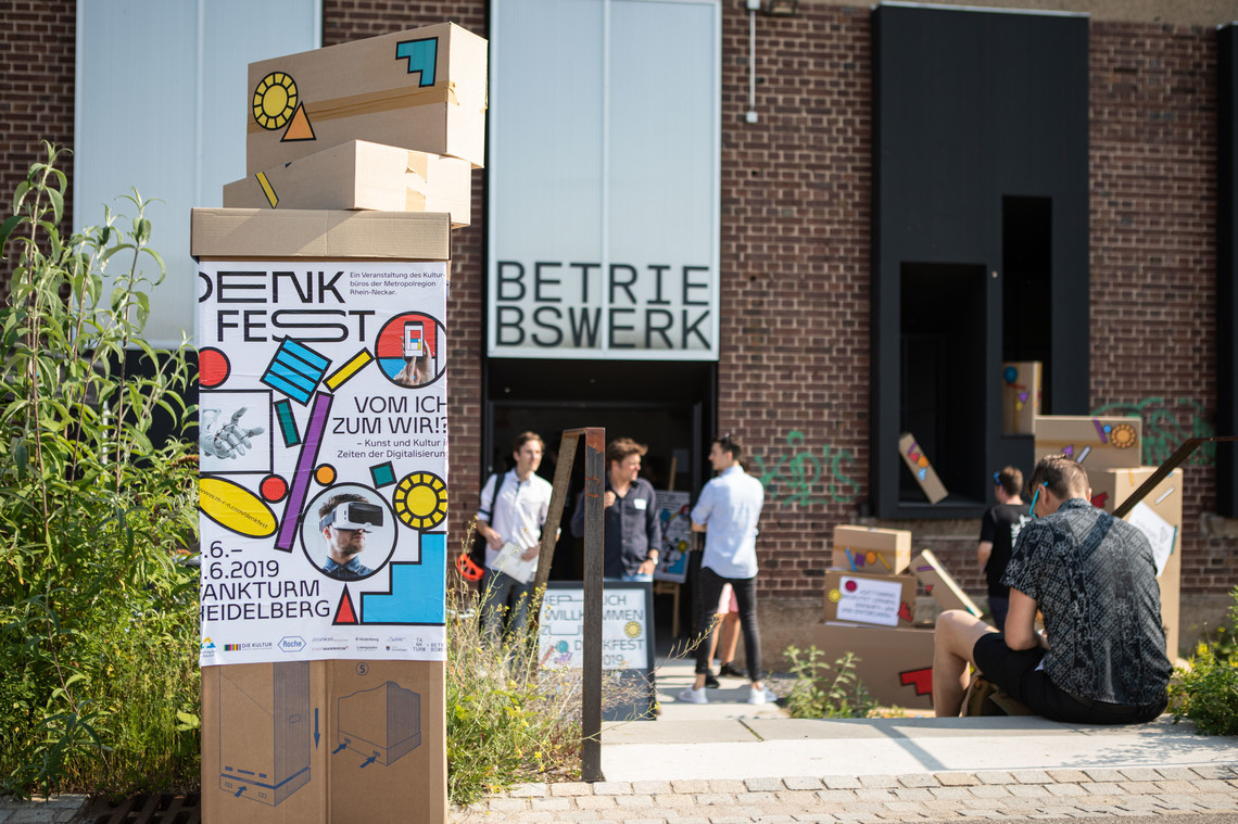Kartons mit Denkfest Schriftzug stehen gestapelt im Vordergrund, im Hintergrund mehrere Personen, die aus einem Gebäude kommen | © Arthur Bauer