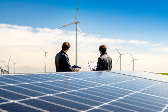 PV-Modul im Vordergrund, dahinter zwei Mitarbeitende mit Tablet und Klemmbrett. Im Hintergrund mehrere Windkraftanlagen | © MRN GmbH