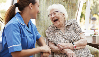 Seniorin lacht mit Pflegerin auf einer Parkbank vor einem Baum. | © Adobe Stock / Monkey Business