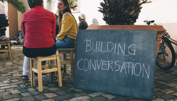 BUILDING CONVERSATION in Neustadt an der Weinstraße beim Demokratiefestival HAMBACH! im September 2018 | © Sarah Hähnle Zwei sitzende Personen im Gespräch, Abendlicht auf einem öffentlichen Platz, Palmentopf in Hintergrund, eine mit Kreide beschriebene Tafel zeigt die Worte "Building Conversation" | © Sarah Hähnle