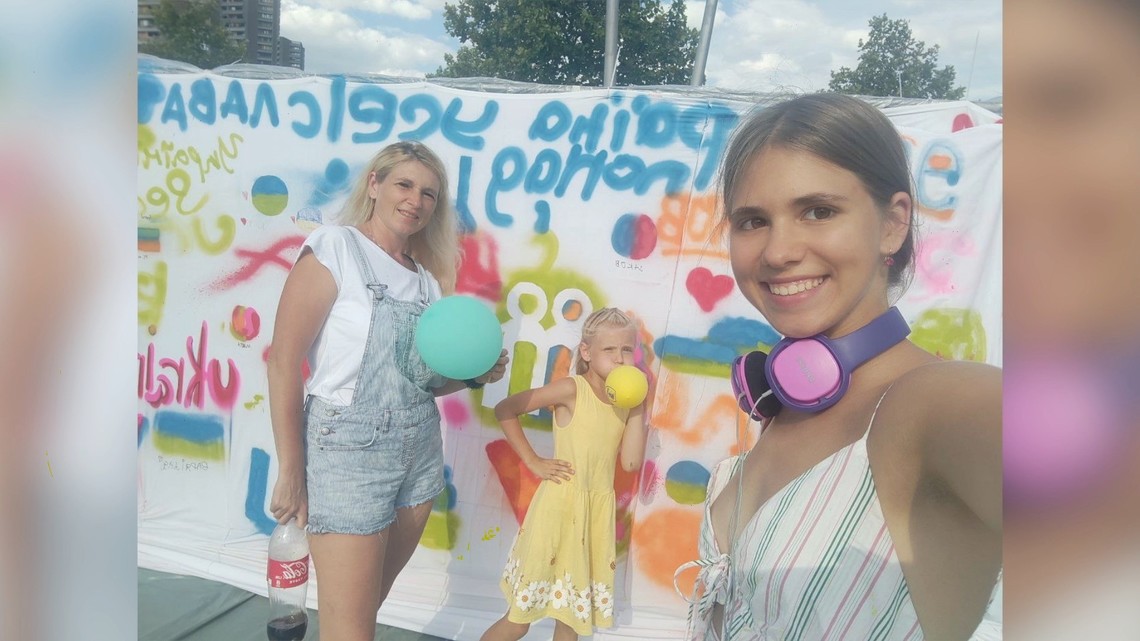 Selfie eines Mädchens mit ihrer Mutter und kleinen Schwester. Im Hintergrund weißer Stoff der mit bunten Texten auf ukrainisch bemalt wurde | © Girls Go Movie / Meine Geschichte